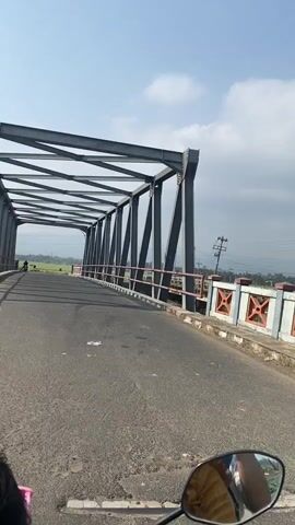 A first-person perspective video shows a person riding a motorcycle across a metal truss bridge. The road surface is asphalt. The bridge has a black metal structure with diagonal beams and a lighter colored railing. On the left side of the bridge, there is a green field. On the right side, there are some buildings and power lines visible. The sky is blue with some white clouds. The motorcycle rider's reflection is visible in the rearview mirror.