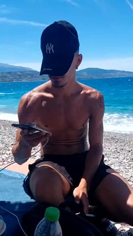 A shirtless person wearing a black baseball cap sits on a beach, looking at their phone. The person is wearing black shorts and has tattoos on their arm. They are sitting on a blue towel next to a bottle of water. The ocean is in the background with waves crashing on the shore. The sky is blue with some clouds.