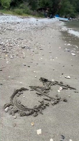 The video begins with a close-up of sand on a beach. A crude drawing of a handicap symbol is visible in the sand, surrounded by small pebbles. As the camera zooms out and pans upward, it reveals a calm, clear body of water with small waves gently lapping the shore. In the distance, a rocky, mountainous landscape is partially obscured by low-hanging clouds or fog. The shoreline is lined with trees and vegetation. A small, light-blue boat is docked on the shore. Two people are swimming in the water near a white yacht in the distance. The overall tone of the video is peaceful and serene, emphasizing the natural beauty of the beach and surrounding landscape.