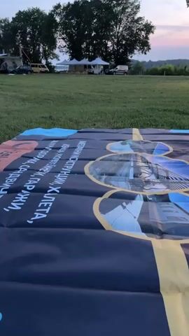 A woman stands in a grassy field next to a large banner. In the background are trees, some buildings, and what appears to be an outdoor seating area with tables and umbrellas. The sky is a light blue, indicating either early morning or late evening. The camera pans slowly to the right, showing more of the field and the distant structures.