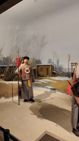 The video displays a museum exhibit featuring life-size mannequins in historical Chinese attire. The scene depicts a snowy, outdoor setting with a backdrop of a small building and bare trees. In the foreground, two male mannequins are positioned near a brown horse. The mannequin on the left is dressed in a long grey robe, a vest, and a black hat with a red pom-pom. He holds a spear with a red tassel. The mannequin on the right wears a dark blue robe, a red hat, and a yellow sash. He appears to be interacting with the horse, possibly adjusting its bridle or reins, while the other mannequin looks on. A second mannequin, similar in dress to the one on the right, is partially visible on the far right of the frame, also near the horse. Another mannequin, standing and holding a spear with a red tassel, is on the far left of the frame. A red banner with Chinese characters is visible in the background. The overall impression is a historical diorama representing a scene from the past.