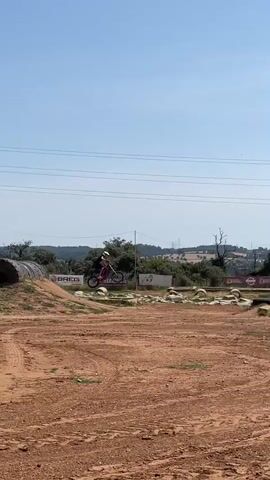A person rides a bicycle over a dirt course. The person is wearing a helmet and a black outfit. They are riding a bicycle that appears to be dark or purple in color. The course is made of red dirt and has various obstacles such as rocks, tires, and small hills. Banners with the words “BREG” and “SEAKING.ES” can be seen on the edge of the course. In the background, there are green trees and rolling hills under a clear blue sky. Power lines are visible overhead.