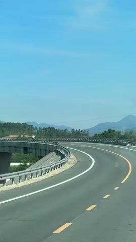 The video shows a road curving to the right with mountains in the background under a clear blue sky. There is a bridge to the left of the road with guardrails along its edge. The road is paved and has white and yellow lane markings. Trees and vegetation are visible on the hillsides. A single power line tower is also visible on one of the hills. The perspective suggests the video is being recorded from a moving vehicle.