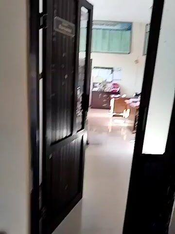 The video shows an interior office space. The camera appears to be positioned just inside a doorway, looking into the room. On the left, a dark wooden door is ajar, revealing part of an office chair and filing cabinets. The room itself has light-colored tiled flooring and light-colored walls. Two large charts or posters with rows of text and numbers are mounted on the far wall. In the center of the room, there are two desks, each with papers, folders, and other office supplies. A dark brown filing cabinet is visible behind the desk on the left. The lighting suggests it is daytime, with natural light coming from unseen windows or the doorway. The scene is static, with no people visible.