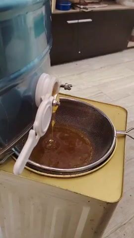 A close-up shot shows honey being poured from a blue extractor into a metal strainer. The strainer is positioned above a golden container, and the honey drips through the mesh. The extractor has a white spigot with a handle, and a metallic valve. The background is slightly blurred, showing a wooden floor and a dark cabinet. The lighting is indoor and appears to be artificial. The video appears to be a still frame, with no discernible movement.