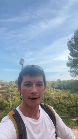 The video begins with a close-up selfie of an Asian man with short dark hair, sunglasses perched on his head, and a white t-shirt. He is wearing a backpack with orange straps. The man is smiling and making a peace sign with his right hand. In the background, a large stone aqueduct with numerous arches stretches across a hillside covered in green vegetation. The sky is blue with scattered white clouds. The camera pans slightly to the left, revealing more of the aqueduct and the surrounding landscape. The man continues to smile and interact with the camera, occasionally tilting his head. The lighting suggests it is daytime, likely late afternoon or early morning due to the warm tones. The environment appears to be outdoors, possibly a tourist attraction or historical site.