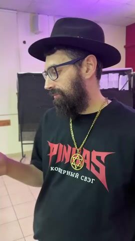 A bearded man wearing glasses and a black fedora is shown from the chest up in an indoor setting. He has a gold-colored chain with a Star of David pendant around his neck. He is wearing a black t-shirt with the word "PINHAS" in red, stylized letters, and "КОШЕРНЫЙ СВЭГ" (Kosher Swag) in Cyrillic beneath it. The man is looking slightly to his left and appears to be speaking, making small hand gestures. The background features a white wall with a speaker on the left, a black backdrop, and a red wall section to the right. The room has light-colored tiled flooring and a purplish ambient light. This scene takes place in Rehovot, Israel.