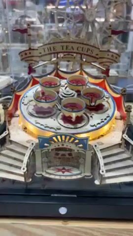 The video shows a miniature model of "The Tea Cups" carnival ride under a clear plastic display case. The model features six tea cups painted with red and white details on a white circular platform with yellow LED lighting. The platform rotates. Above the tea cups is a sign reading "The Tea Cups," decorated with flags and stars. The base of the model is designed with steps leading up to an archway. Behind the tea cups is another miniature model of a Ferris wheel. The model sits on a black base with a white button, presumably for operation. The video is shot indoors, showing other displays in the background.
