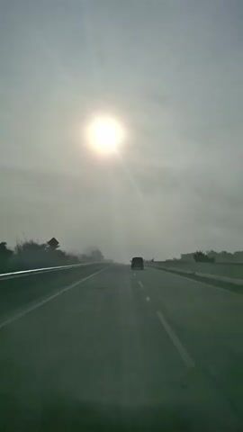 A vehicle drives on a highway under a bright sun and a hazy sky. The sun is a large, bright orb in the upper center of the frame, casting rays downwards. The sky is mostly overcast with a light gray haze. The highway stretches forward, with two lanes visible in each direction. A white dashed line separates the lanes. On the left side of the highway, there is a guardrail and some sparse vegetation. In the distance, there are trees and hills. A dark-colored vehicle is visible ahead in the right lane, moving in the same direction. The overall scene suggests a dusty or foggy environment.