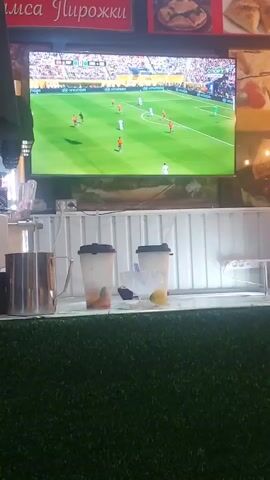The video shows a large television screen displaying a soccer match. The screen is mounted above a counter with several objects on it, including two to-go cups with lids, a metal pitcher, and some scattered items. The counter is covered with what appears to be artificial grass. In the background, there are signs with text in a foreign language and images of food, possibly baked goods. The overall environment appears to be an indoor establishment, possibly a cafe or food stall, where patrons can watch sports while consuming food and drinks.