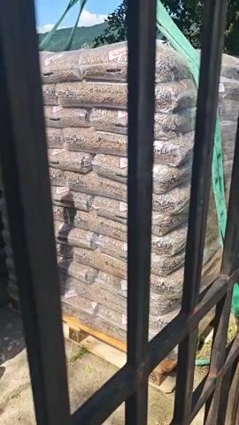 The video begins with a view through a dark metal fence of a stack of bags, possibly containing wood pellets, being lifted by a green strap. A person wearing an orange t-shirt and denim shorts is visible, holding the strap. The camera then pans to the right, showing a rusted metal mailbox attached to a black pole, with a wire fence in the background. The mailbox has a slot for mail and a lock. The video ends on this close-up of the mailbox.