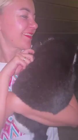 A woman with blonde hair, red lipstick, and a white t-shirt is holding a black and white cat. She is smiling at the camera. The cat is resting on her shoulder. The background is blurry, but it appears to be indoors, possibly a kitchen or dining area. There are glimpses of furniture and a wall decoration.