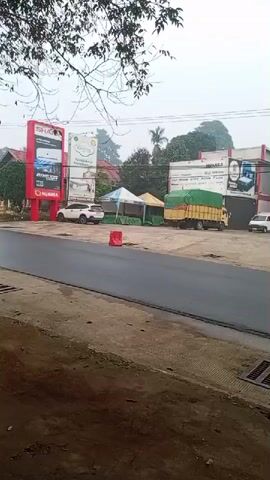 The video shows a street view in front of a building with multiple businesses. A white SUV and a yellow truck covered with a green tarp are parked in front of the buildings. A small white truck is also visible. There are several tents set up in front of the buildings. The road in front of the buildings is newly paved and appears to be a fresh asphalt surface. The overall environment appears to be a commercial or industrial area with some vegetation visible in the background. The sky is overcast.