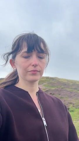 A medium shot video features a woman with dark hair and bangs, wearing a dark maroon zippered jacket and earrings. The background is an outdoor, hilly landscape with sparse purple vegetation and a cloudy sky. The woman's head moves slightly, at times looking directly at the camera with a neutral or slightly somber expression, and at other times looking to the side. The video captures the natural movement of the woman's head and hair in what appears to be a windy environment.