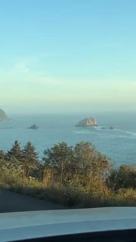 The video shows a view from a vehicle looking out at the ocean. The ocean is a deep blue and there are several rock formations in the water. Trees and vegetation are visible on the shoreline in the foreground. The sky is clear and blue, with a slight haze near the horizon. The lighting suggests it is late afternoon or early evening. There are no people visible in the video.