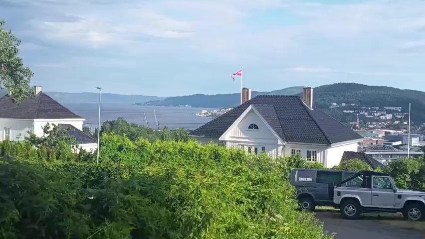 The media file displays a scenic view of a coastal town with hilly terrain, likely Drammen, Norway. The time of day appears to be late afternoon or early evening, with soft, natural lighting suggesting fair weather.

The foreground is dominated by lush green foliage and a paved driveway. Parked on the driveway are two vehicles: a dark-colored utility vehicle with a gray canopy, and a silver, open-top four-wheel-drive vehicle. The utility vehicle has "ROCKTON" written on its side in white letters.

In the mid-ground, a white two-story house with a dark roof stands prominently. A Norwegian flag flies from a chimney on the house. Further into the scene, the town unfolds with buildings scattered across the hillsides. A body of water, likely a fjord or bay, stretches into the distance, dotted with ships. A bridge is visible spanning the water.

A young man, dressed in a striped short-sleeved shirt and shorts, walks along the paved area in the mid-ground, casting a shadow. He appears to be casually strolling, with no indication of urgency or strong emotion.

A street sign with Norwegian text is visible among the trees. The text on the sign appears to be "Strømsveien" and "Fjordbuergate".

The overall impression is one of a peaceful, residential area with a beautiful natural backdrop. The clear sky and abundant greenery suggest a pleasant summer day.