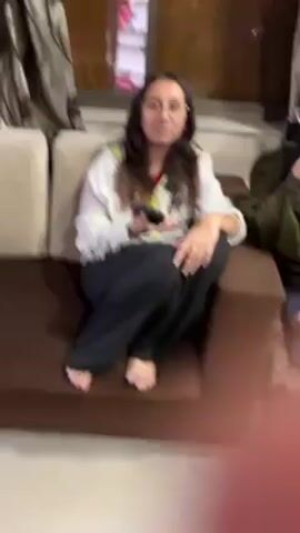 The video shows several women and a child dancing in what appears to be a home setting. Initially, two girls are sitting, one on a sofa and the other on a stool. Then, the camera pans to show a group dancing, including a woman in a purple patterned outfit and another in a red salwar kameez. A child in pajamas is also dancing. A woman in a sari joins the group. The background includes furniture and home decor. Music is playing.