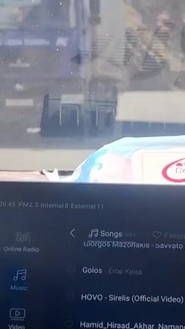 A video taken from inside a car shows a drive on a mountain road. The viewpoint is from behind the dashboard, with a package of baby wipes visible in the foreground. The car is traveling on a winding, paved road with steep, rocky mountain slopes on either side. There are wooden utility poles lining the side of the road, some adorned with colorful markings. The weather appears to be clear and sunny. The car is climbing in altitude, indicated by the phone screen showing an elevation of 2618 meters and a temperature of -3 degrees Celsius. Music is playing on the phone.
