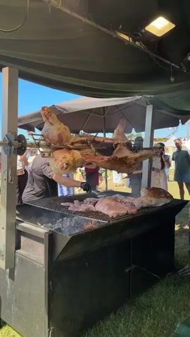 This video shows a roasting spit with several large cuts of meat, possibly lamb or pig, suspended above a charcoal grill. Smaller cuts of meat are being grilled on the grate below. A man in a black shirt stands to the left of the grill, and other people are visible in the background, some standing under tents and others walking through a grassy area. The scene appears to be an outdoor food stall or festival, with bright sunlight and a blue sky overhead. The spit is being turned by a mechanical device. Some of the meat appears to be partially cooked, with browning and charring evident. There is also steam rising from the grill. The overall impression is of a busy, outdoor cooking event.