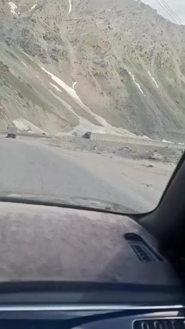 The video shows a view from inside a car driving on a road through a mountainous region. The road is gray asphalt, and there's a truck ahead. The mountains are rocky with patches of snow, and the sky is overcast. A man with a shovel is visible next to a building at the side of the road. The car's dashboard is visible in the foreground, slightly dusty. The overall impression is of a desolate and remote landscape.