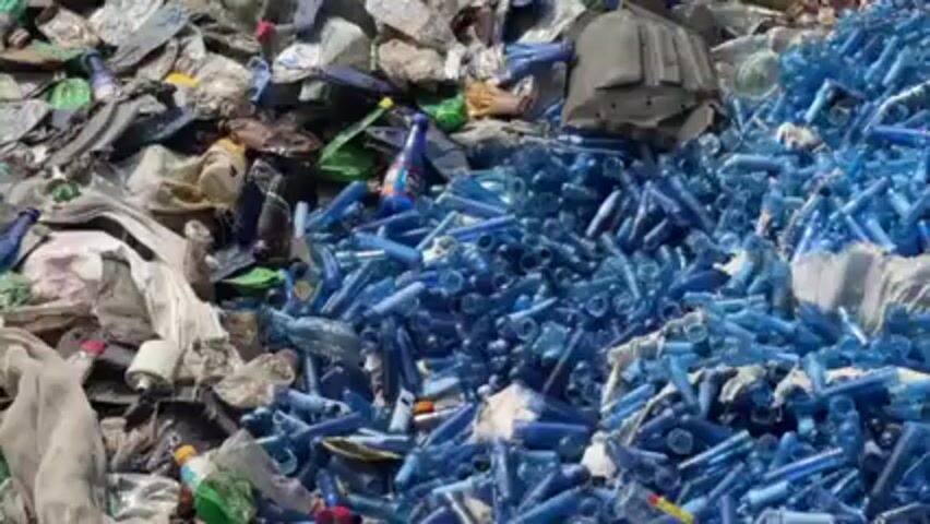 The image shows a large pile of mixed plastic waste. The waste is composed of various types of plastic bottles, containers, and other plastic materials, with a large proportion of it being blue-colored preform bottles. A blue bucket sits on top. The overall appearance indicates a recycling or waste disposal site.