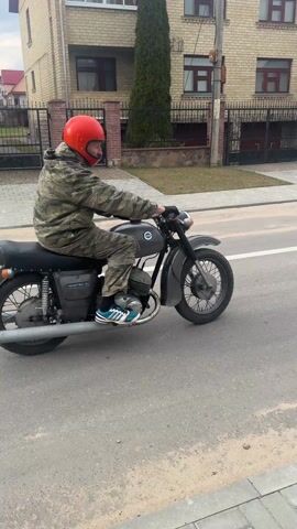 A person wearing a camouflage suit and a red helmet drives a grey motorcycle along a street, then turns onto a sidewalk of grey rectangular stones. There is a parked silver car and several buildings in the background. The sky is overcast.