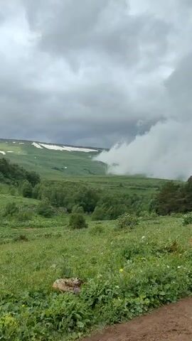 A lush green meadow slopes gently downwards towards a dense forest. Beyond the forest, a vast expanse of green land stretches out, dotted with patches of snow and shrouded in swirling mist. The sky above is heavy with dark, dramatic clouds, suggesting an impending storm. The foreground is filled with thick green grass and wildflowers, with a dirt path visible on the right side of the frame. The overall impression is one of raw, untamed natural beauty, with a sense of the elements at play. There are no people or animals visible in the video.
