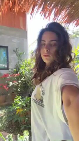 The video shows a woman standing in a garden, under a thatched roof. She wears a white t-shirt and has long, wavy brown hair. She raises her hand to her head, revealing a tattoo on her lower back.  In the background are flowers, cacti, and a building with a window.