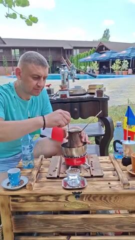 A man in a teal t-shirt and dark shorts is seated at a wooden table outdoors. He is stirring a copper pot on a small red stove with a metal spoon. In front of him on the table are a white cup with blue dots, a bottle of water, and a small stack of coffee beans. To his right is a small flag, a Bialetti espresso maker, and another cup. In the background, there is a swimming pool, a building with a brown exterior, and outdoor seating with umbrellas. The scene is illuminated by daylight.