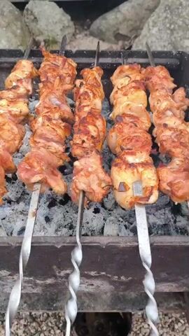 Here's a detailed description of the video:

The video shows a close-up view of several skewers of marinated and partially grilled meat over a charcoal grill. The skewers are made of silver metal and have a twisted design on the handle end. The meat appears to be chicken or pork, cut into roughly equal pieces and threaded onto the skewers. The meat is browned and shows some charring in spots.

The grill is made of dark metal and sits over a bed of glowing white-hot charcoal. There is a light amount of smoke rising from the cooking meat. The grill is situated outdoors, with light-colored gravel visible around its base. In the background, there are a few large, grey rocks.

There is a small child speaking in the background.