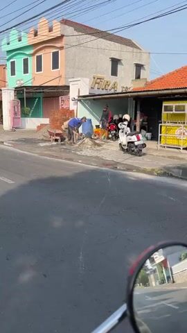 The video shows a street scene with a building under construction. A man in a blue shirt is shoveling cement from a pile into a wheelbarrow. Two other individuals are standing nearby, one of whom appears to be operating a concrete mixer. A white scooter is parked on the side of the road. The building has a colorful facade, with sections painted mint green, pink, and gray. A sign above one of the shops reads "FIRA." The sky is clear and the sun is shining. The video is filmed from the perspective of someone on a moving vehicle, likely a motorcycle, as indicated by the reflection in the rearview mirror.