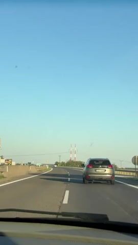 A first-person perspective video captures a drive on a highway under a clear blue sky. The camera is positioned inside a car, showing the dashboard and windshield. A dark-colored SUV is driving in front and to the right of the camera car, on the same lane. Further down the highway, more vehicles are visible. On the left side of the road, a concrete barrier separates the highway from a field. In the distance, two large, red and white electrical pylons stand tall against the horizon, with a few smaller utility poles scattered along the landscape. The sun is setting, casting a warm glow and reflecting in the car's side mirror.