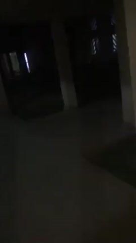 The video shows an exterior shot at night, likely taken from a mobile device. The scene appears to be the exterior of a building, possibly a one or two story structure, with lights illuminating the windows or an open walkway. The camera seems to move erratically, initially showing what may be an overhang or covered walkway before panning out to reveal the building. There is a dark, grassy area in the foreground. The overall lighting is poor, making it difficult to discern specific details. There are no discernible people or interactions in the shot.