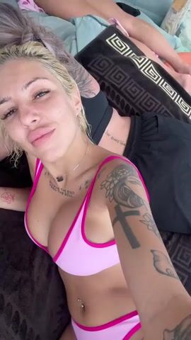 Two individuals are lying down on what appears to be a beach or outdoor seating area. The foreground subject, a woman with blonde hair and numerous tattoos, is looking directly at the camera. She is wearing a pink bikini and has a navel piercing. Other tattoos are visible on her arms and chest.

Behind her, another individual with greyish-blonde hair pulled back in a bun is also lying down, looking away from the camera and holding a smartphone. This person is wearing sunglasses and has visible tattoos on their arm. A third person is partially visible in the background, also lying down.

Objects scattered around include a pink mesh bag filled with items, a phone, a water bottle, and what looks like a towel with a patterned edge. The scene suggests a relaxed, possibly vacation setting. The lighting indicates it could be daytime. There is some visible text on the foreground subject's chest that reads "NOTHING IS AS IT SEEMS." and "THE EGO."