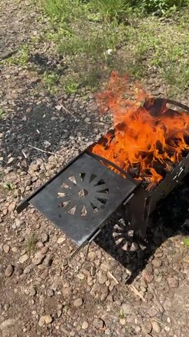 A portable metal barbecue grill with an open flame is situated on a rocky, outdoor surface adjacent to a body of water and grassy vegetation. The grill is black, with a decorative circular pattern on its side and a flat, hinged metal surface on its left. Flames engulf the contents of the grill, emitting orange and yellow light. The surrounding ground is covered with small stones and scattered dry leaves. Lush green grass and foliage are visible in the background, with a calm body of water bordering the scene on the left. The lighting suggests it is daytime, likely sunny. There are no people or animals visible in the frame.
