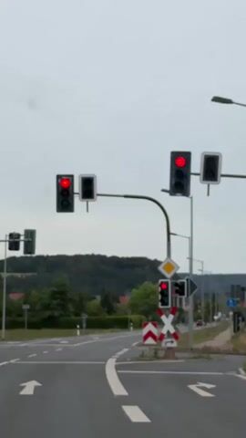 The video shows a view from a vehicle driving on a road. The camera is focused on traffic lights and road signs. Multiple traffic lights are visible, all displaying a red signal, indicating a stop. There are also signs indicating a railway crossing. The environment appears to be a suburban or rural area with trees and hills in the background. The sky is overcast.