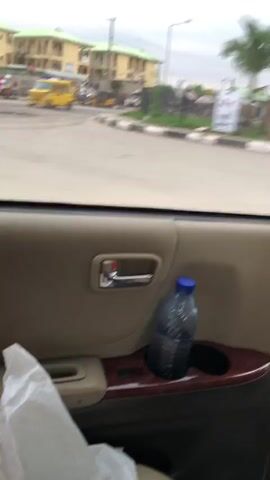 The video shows a view from inside a car, looking out the window. The focus is on a large, abstract sculpture of a person, partially obscured by trees. In the foreground of the car's interior, there's a door handle and a water bottle in a cup holder. Outside, there's a street with a few motorcycles and a white car with "DRIVING ACADEMY" written on it, along with the number 122. The sky is overcast, and buildings and palm trees are visible in the background. The car appears to be moving.