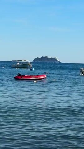 A wide shot shows a calm, blue ocean under a clear sky. Several boats are visible on the water. In the foreground, a red inflatable dinghy floats with a rope attached. To the right, a white motorboat with a canopy is moving slowly and appears to have several people on board. In the middle ground, a white boat with a canopy is anchored. In the background, a large white cruise ship is visible on the horizon. The water is gently rippling.