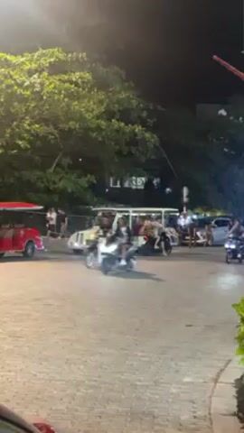 At night, a street scene unfolds with a collection of vehicles and pedestrians. Several scooters, each with a driver, maneuver around the paved intersection. Among the vehicles is a white, elongated golf cart, open on the sides and occupied by several people. Further back, a red tuk-tuk-like vehicle is visible. Lush green trees overhang the street, and the lights illuminate the scene. The road is paved with patterned stone blocks.