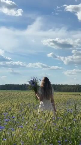 A young woman with long blonde hair, wearing a white dress, walks through a field of green and yellow wheat. She is carrying a bouquet of blue flowers. The sky is blue with white, fluffy clouds. In the background, there is a line of trees. The camera is positioned behind the woman, capturing her from the back as she moves away from the viewer. The lighting suggests it is daytime, possibly late afternoon, with the sun illuminating the scene.