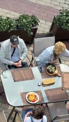 A high-angle shot shows three people seated at an outdoor cafe table. A man in a striped shirt and a "DAD" cap sits to the left. A woman in a light blue shirt and straw hat is seated across from him. A child in a denim overall dress is at the end of the table. Two plates of food are visible: one with sausage, beans, and fried items, and another with food in a small frying pan. Two phones are placed on the table. There are potted plants near the table. The ground is made of bricks. The people seem to be waiting to eat.