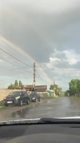The video, shot from inside a vehicle, shows a street scene after a rain shower. A double rainbow is visible in the sky, arching over houses and trees. The asphalt road is wet and reflects the sky. Two black cars are parked on the left side of the road. Utility poles with numerous wires stretch across the scene. The sky is partly cloudy with patches of blue visible.