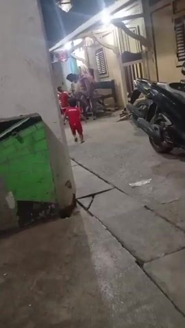 A brief video shows a narrow alleyway at night. The alley is paved with concrete and has a small drainage ditch running along the right side. On the left, a rough concrete wall is visible, with a section painted bright green. Further down the alley, several people are gathered near the entrance of a building. A child in a red shirt walks away from the camera, towards the group. One person in the group appears to be sitting on a wooden bench. A motorcycle is parked on the right side of the alley. Lights are on in the building, illuminating the scene.