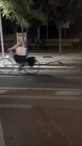 A man on a bicycle rides past a street at night. The man is wearing a blue t-shirt and grey shorts and has white hair. The bicycle has a rear reflector and a front light. There are trees and streetlights in the background. A building with an illuminated window display is visible on the right side of the screen. The window display features plants and a sign that says "Visit PESARO".
