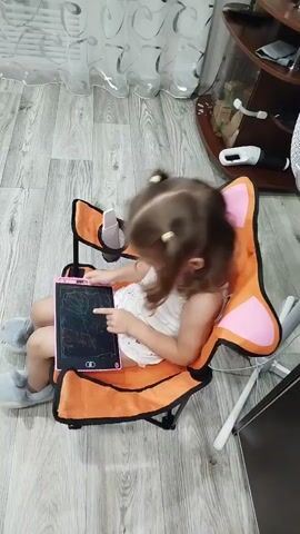 A young girl with blonde hair in pigtails sits in an orange camping chair. She is wearing a white tank top and grey fuzzy slippers. In her hands, she holds a pink electronic drawing tablet, and she is touching the screen with her right index finger. The tablet displays a scribbled drawing in various colors. The girl glances down at the tablet and then looks up, smiling with her mouth open and eyes wide. She gestures with her right hand. The background shows a floor with a wood-grain pattern, a white and grey curtain, and furniture including a dark wood shelf with items on it and a white fan.
