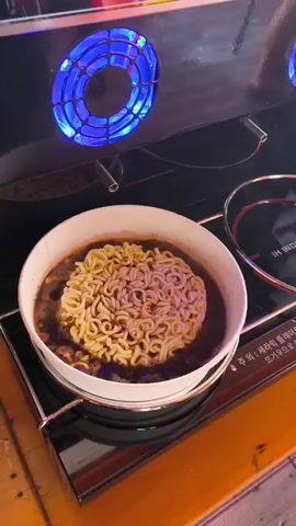 A white bowl of instant ramen noodles sits in dark broth on an induction cooktop. The cooktop has a digital interface with Korean text and icons, indicating different cooking steps and settings. Blue lights surround two circular heating elements on the cooktop. The noodles appear to be partially cooked.