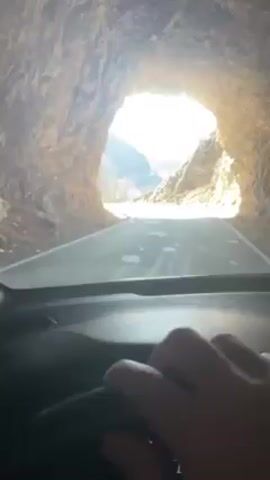 The video shows a first-person view from inside a car driving through a rock tunnel. As the car exits the tunnel, a winding road flanked by steep, rocky cliffs is revealed. In the distance, mountain ranges are visible under a blue sky with scattered clouds. The steering wheel is visible in the lower portion of the frame, with a hand resting on it. The road appears well-maintained with white lines marking the edges.