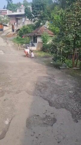 Here is a concise description of the scene: 

The video shows an outdoor setting in what appears to be a rural or semi-rural area. A man is bending over, seemingly picking something up from the ground. A small child in a white dress is standing near him. The man is wearing light-colored pants and a light-colored top. The setting includes a gravelly or dirt road, small buildings with basic architecture, and trees. The scene suggests a peaceful, perhaps everyday moment.