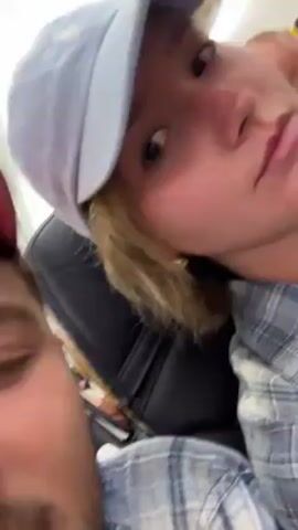 A video shows a man and a woman on what appears to be an airplane. The man has a mustache and is wearing a blue and gray plaid shirt. He appears to be holding the camera. The woman is wearing a white and red baseball cap and a blue and gray plaid shirt. The camera is angled up toward the woman’s face. The man asks the woman how much longer the flight is and she responds “45”.