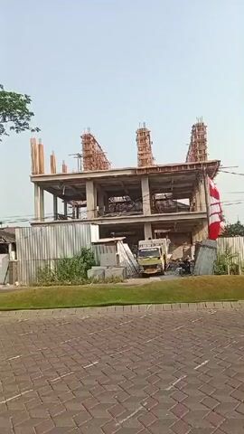 The video captures a construction site on a sunny day. A multi-story building is under construction, with concrete pillars and framework visible on its upper levels. In the foreground, a paved walkway with brick-like patterns leads towards the site. A truck is parked near the building, and various construction materials are scattered around. A motorcycle rider, wearing a helmet and a green vest, passes by on the left side of the frame. The overall environment appears to be an urban or suburban area.
